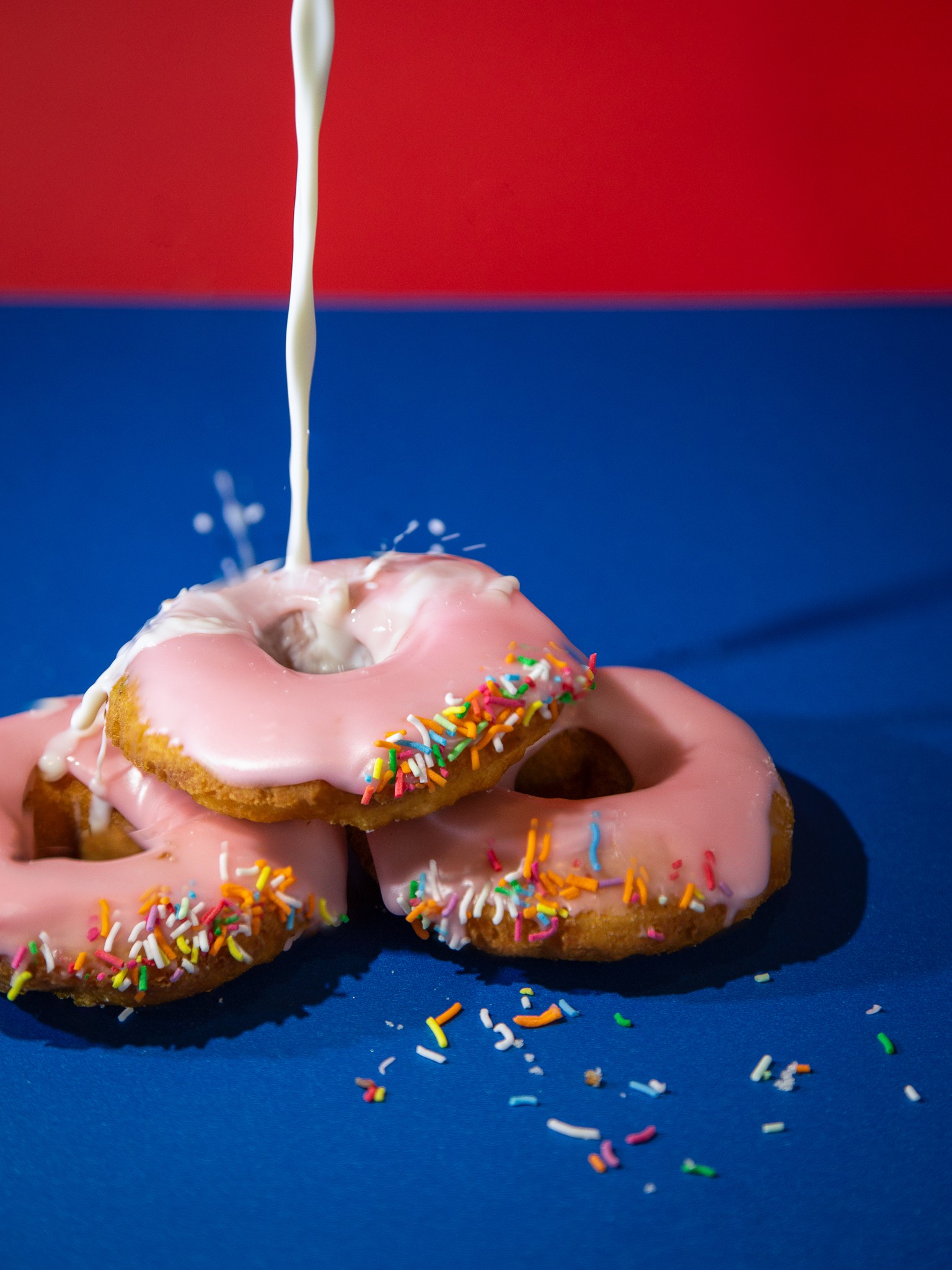 Photograph of fresh milk being poured over pink donuts againt vibrant primary colour background