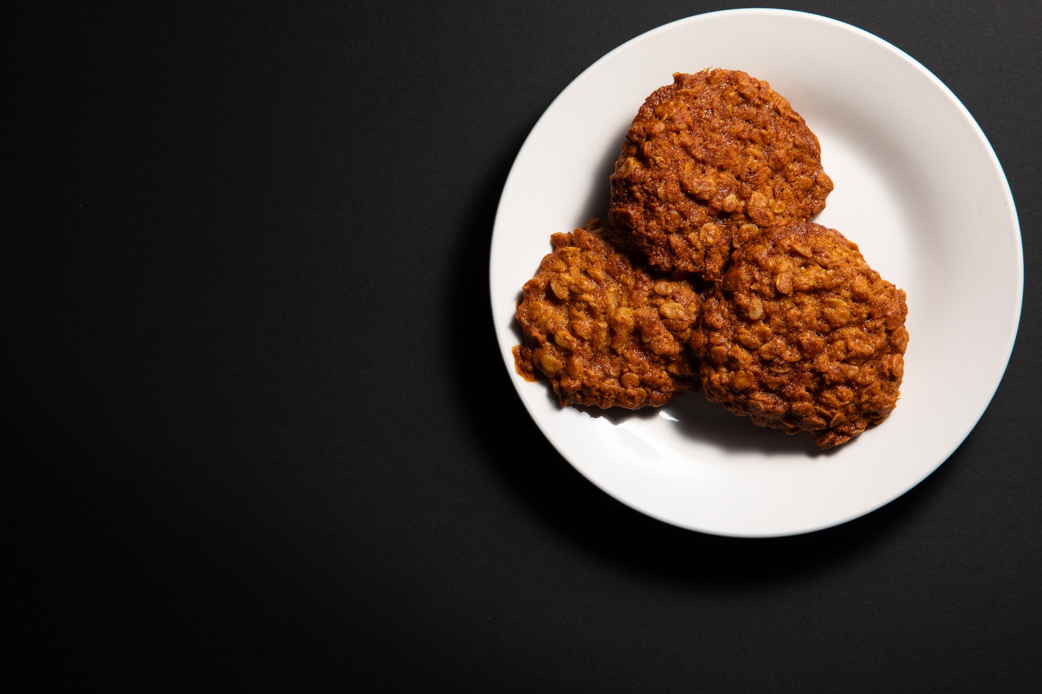 Flat lay food photograph of 3 Anazac biscuits on a white plate with black backround. Negative space for editorial.